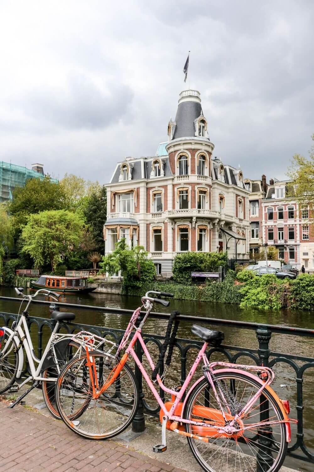 Two bikes, white and pink, lent against a black railing by the river. In the background beyond the river is a dutch style old house with turret and flag. 