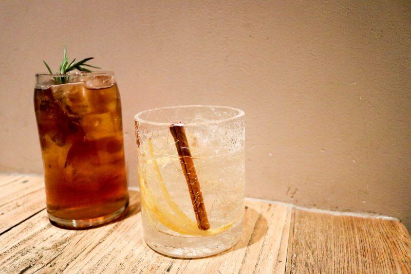 Two cocktails, on a wooden table, one tall and dark garnished with rosemary and one short, clear and garnished with a cinnamon stick. 