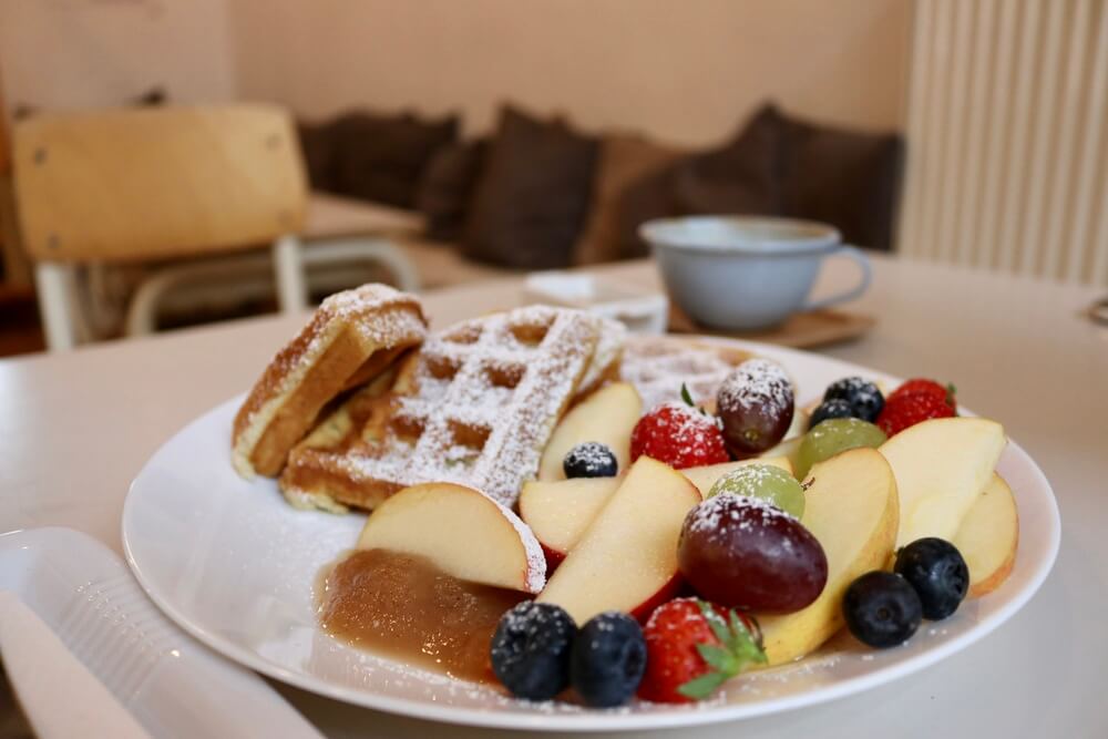 A white plate with icing sugar covered waffle and colourful fresh fruit.