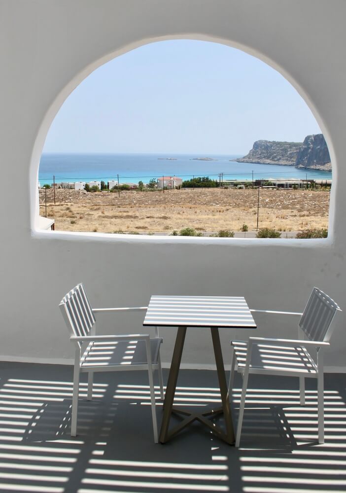 A set of patio chairs either side of a white table, underneath a semi circular window with an ocean view